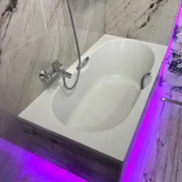 Biała wanna z chromowaną armaturą i oświetleniem LED w łazience wykończonej marmurowymi płytkami i drewnopodobnymi panelami. Nowoczesny design z fioletowym podświetleniem.