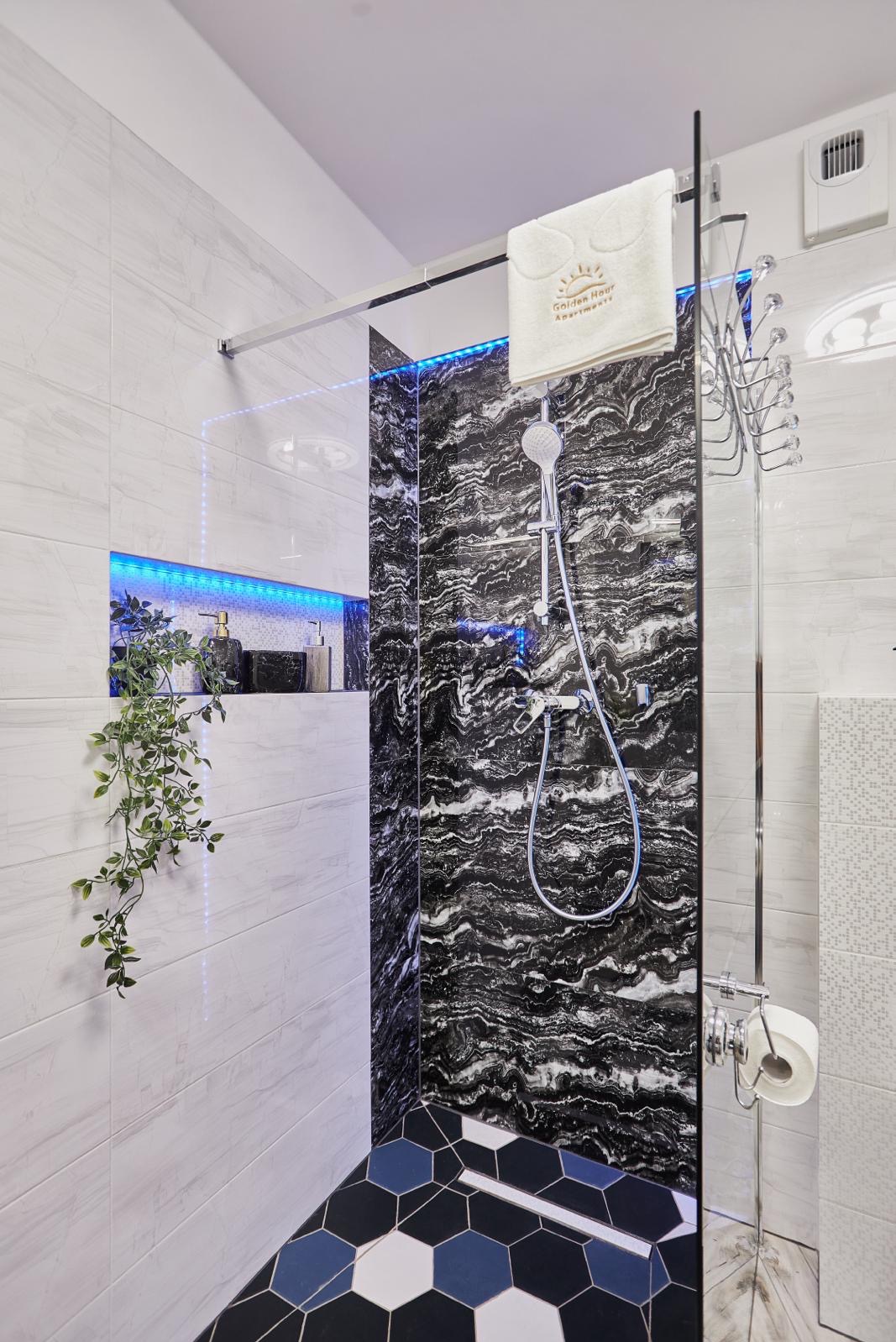 Nowoczesna kabina prysznicowa z czarną marmurową ścianą, oświetleniem LED i heksagonalną podłogą, uzupełniona o rośliny i eleganckie akcesoria łazienkowe.
