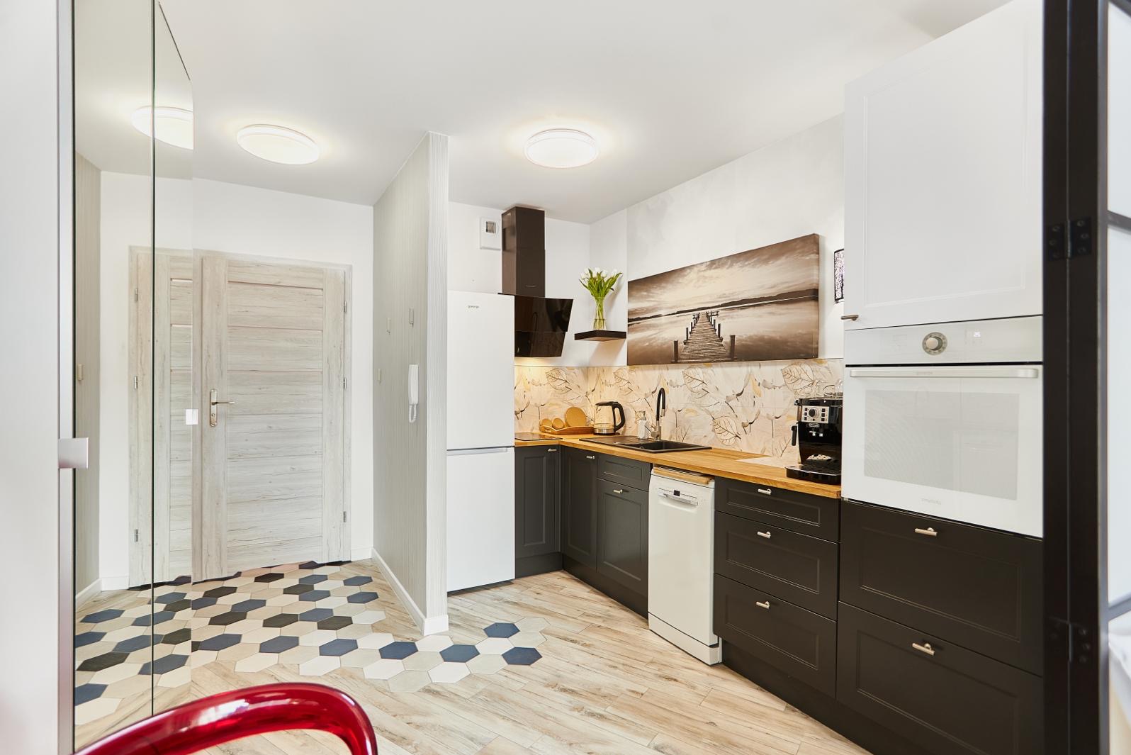 Nowoczesna kuchnia z ciemnymi szafkami, drewnianym blatem i dekoracyjną tapetą. Na podłodze heksagonalne płytki przechodzące w drewnopodobne deski.