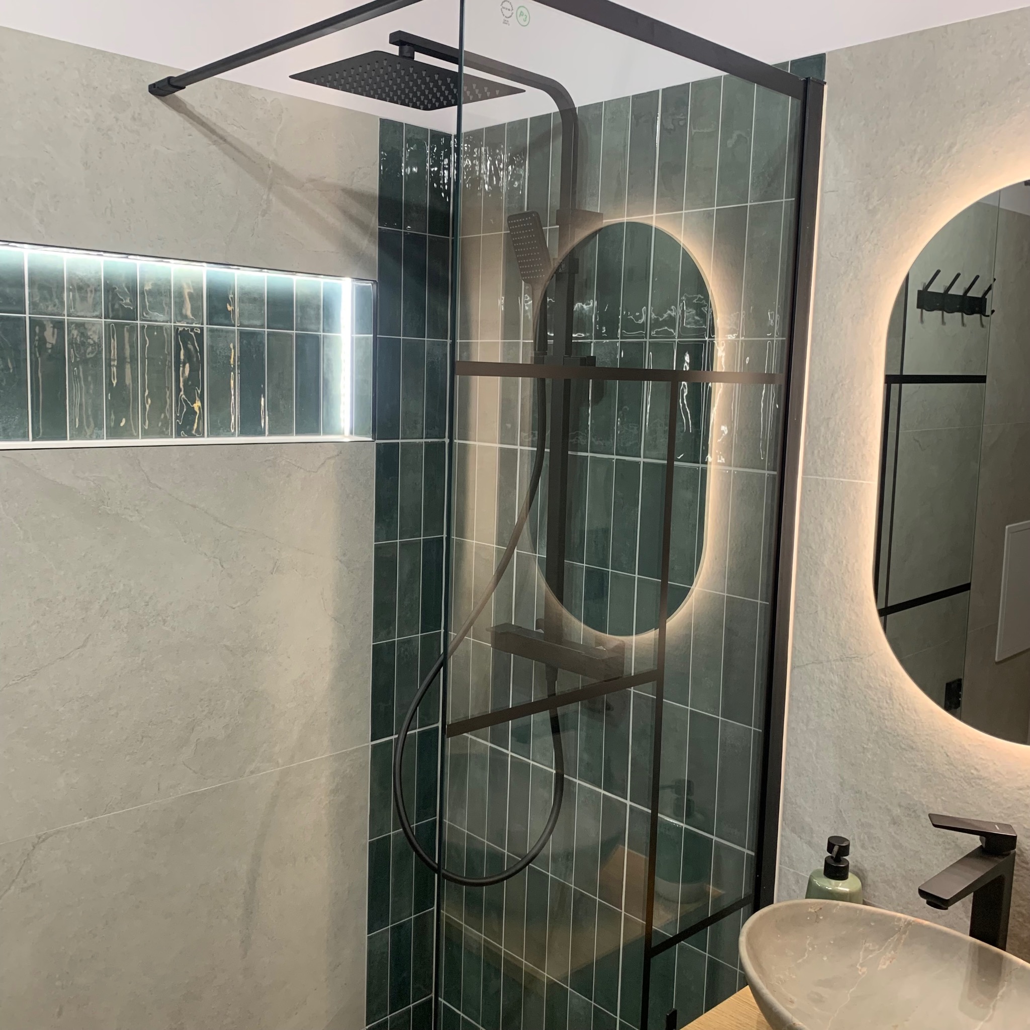 Nowoczesna łazienka z kabiną prysznicową typu walk-in, zielonymi płytkami i okrągłym lustrem z podświetleniem LED. Umywalka nablatowa z czarną armaturą.