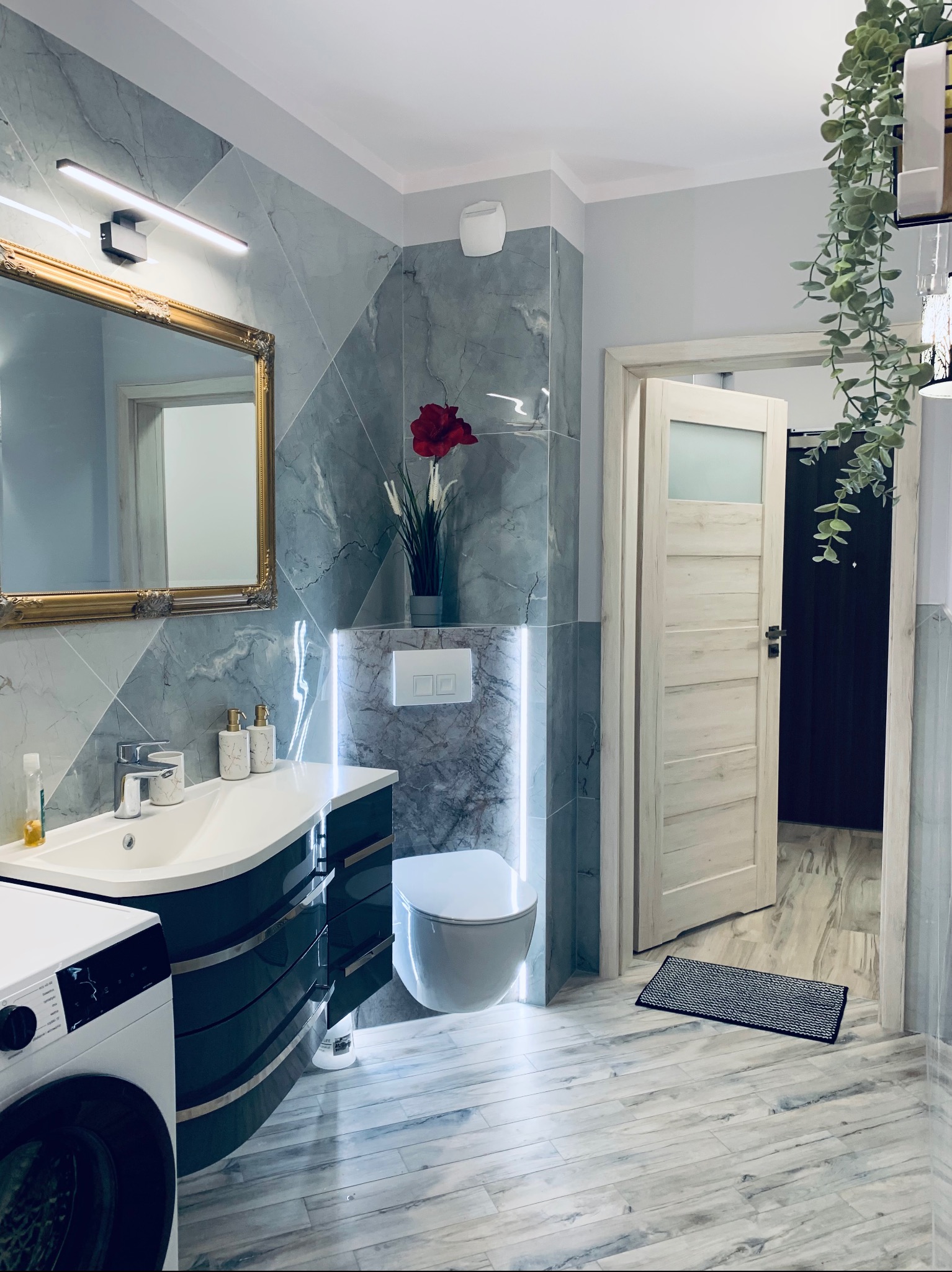 Elegancka łazienka z marmurowymi płytkami, umywalką z szafką w kolorze butelkowej zieleni i złotym lustrem. Oświetlenie LED podkreśla nowoczesny design.