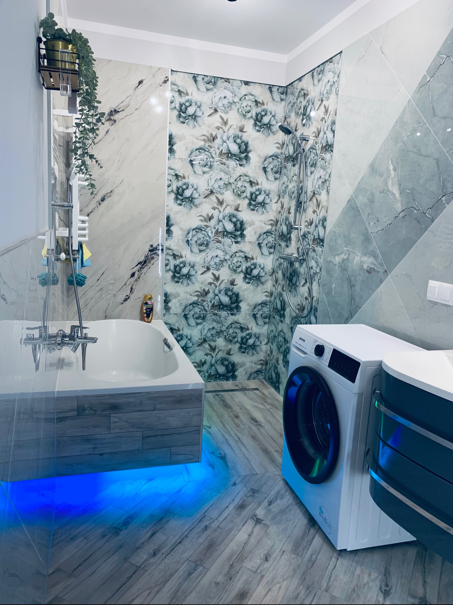 Nowoczesna łazienka z wanną podświetloną na niebiesko, pralką i płytkami w kwiaty. Wykończenie wnętrza z dbałością o detale i oświetlenie.
