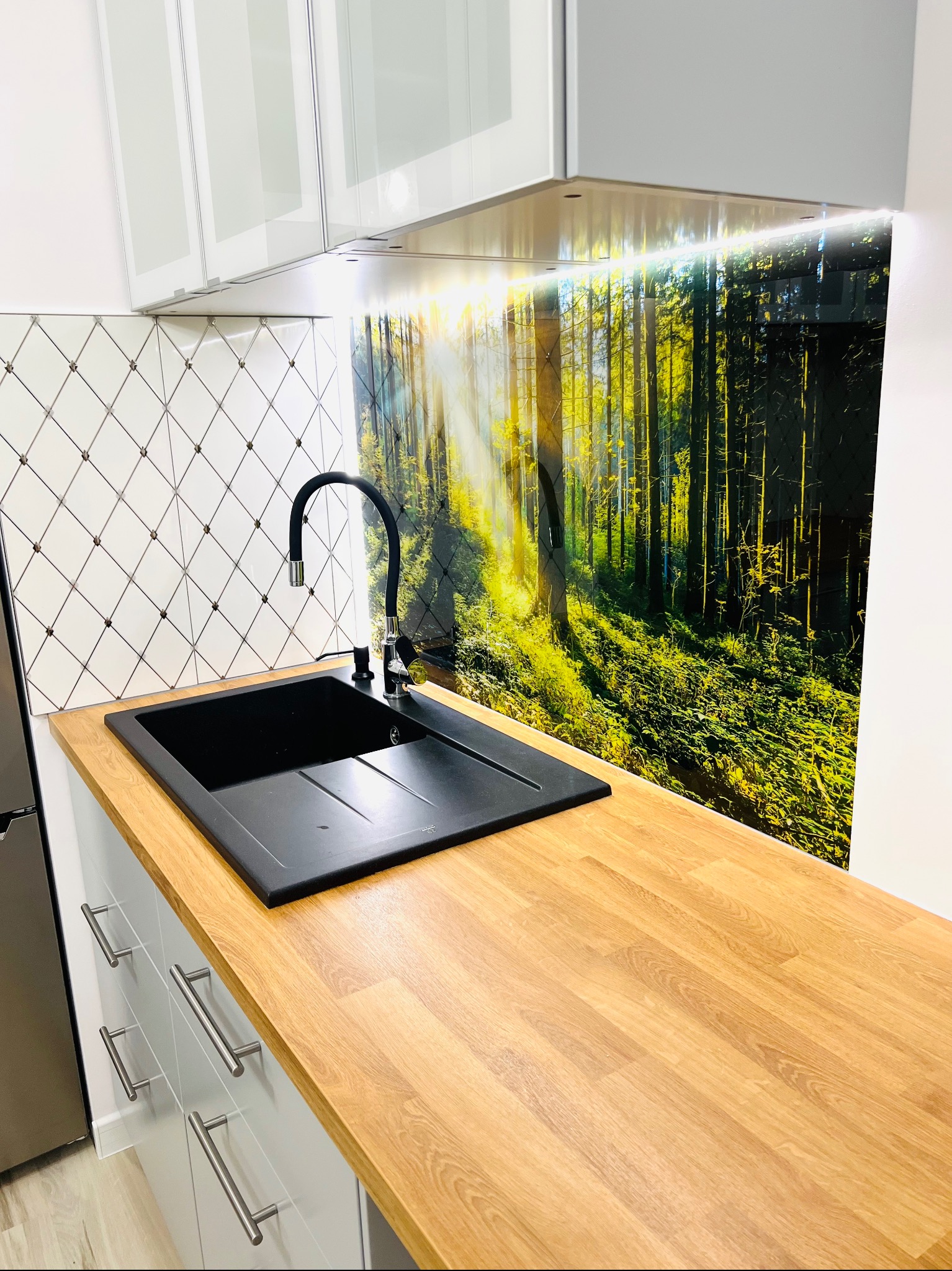 Nowoczesna kuchnia: czarny zlew z baterią, drewniany blat, dekoracyjny panel ścienny z motywem leśnym i białe szafki z chromowanymi uchwytami.