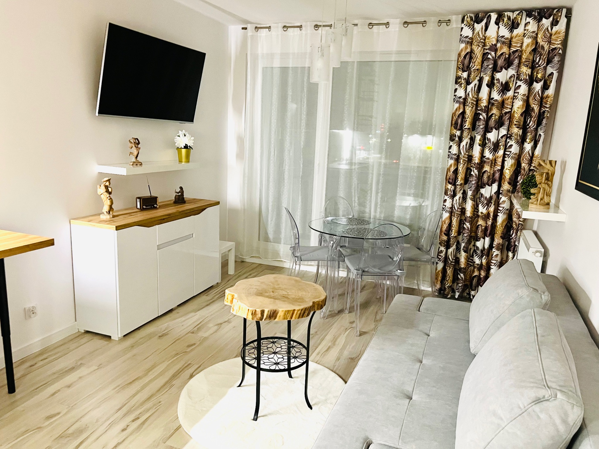 Nowoczesny salon z drewnianą podłogą, szarą sofą i okrągłym stołem z drewnianym blatem. Na ścianie telewizor i dekoracje. Jasne, przestronne wnętrze po wykończeniu.