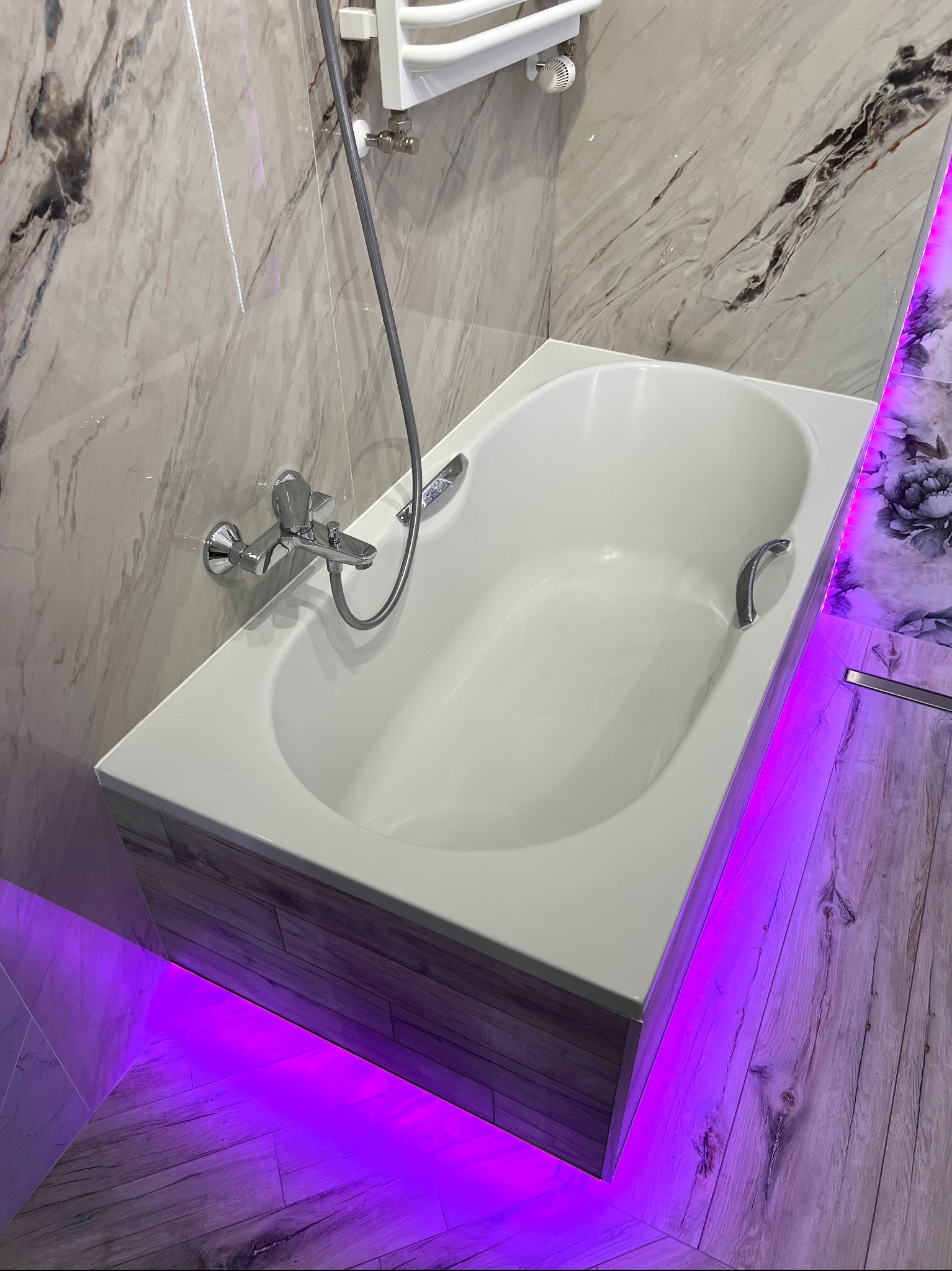 Biała wanna z chromowaną armaturą i oświetleniem LED w łazience wykończonej marmurowymi płytkami i drewnopodobnymi panelami. Nowoczesny design z fioletowym podświetleniem.