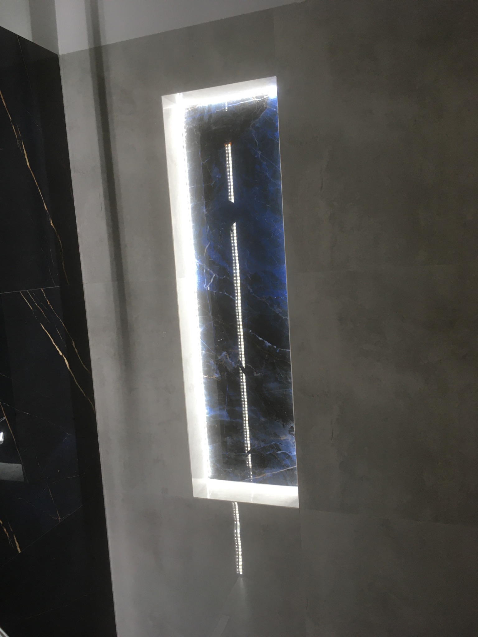 Wnęka z podświetloną płytą imitującą kamień szlachetny, ciemna płytka z żyłkami, szara ściana. Element wykończenia wnętrza łazienki, nowoczesny design.