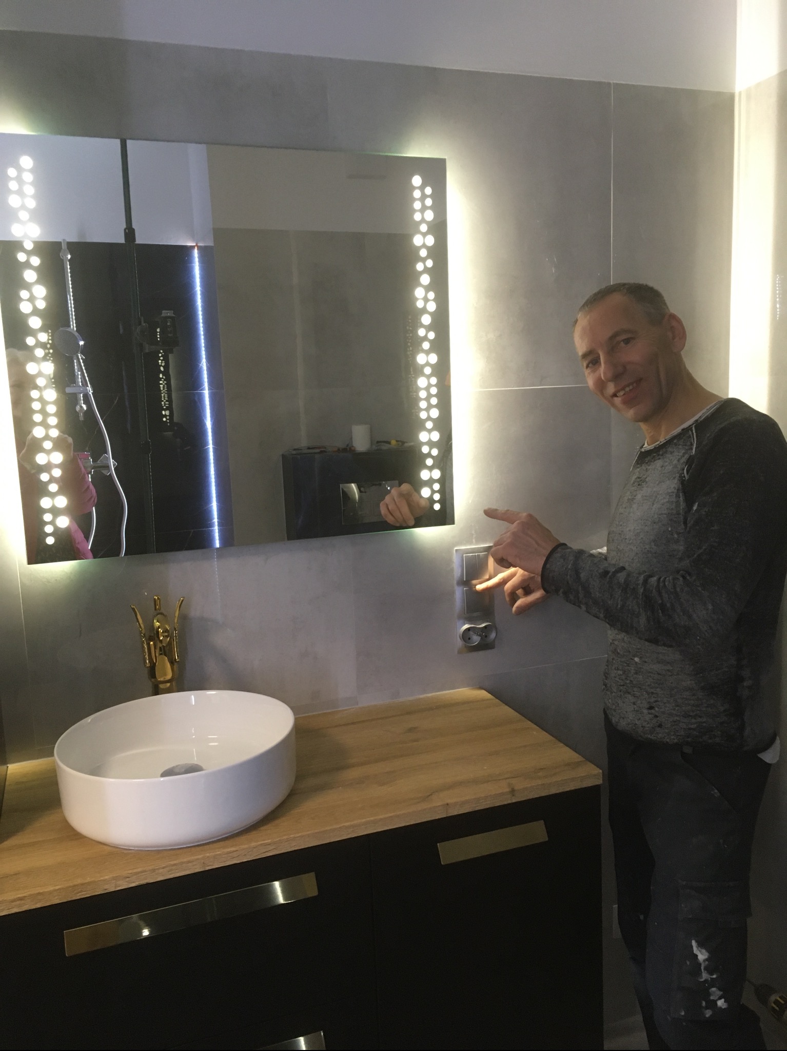 Ukończona łazienka z podświetlanym lustrem, umywalką na szafce z blatem drewnopodobnym. Mężczyzna wskazuje na włącznik światła, szare płytki na ścianach.