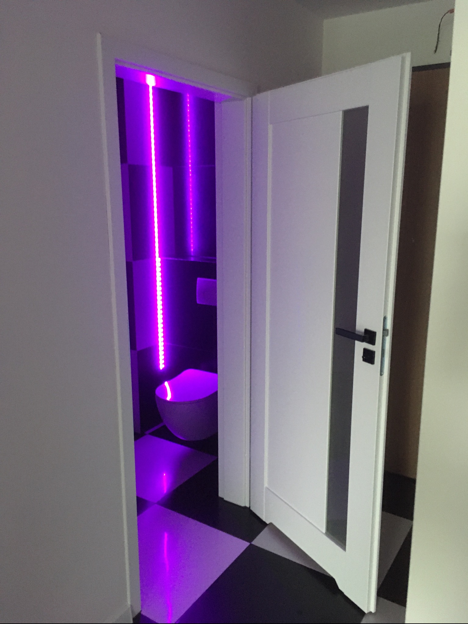 Otwarta biała, nowoczesna wewnętrzna  drzwi z czarną klamką, prowadzące do łazienki z fioletowym oświetleniem LED i wzorem szachownicy na podłodze.