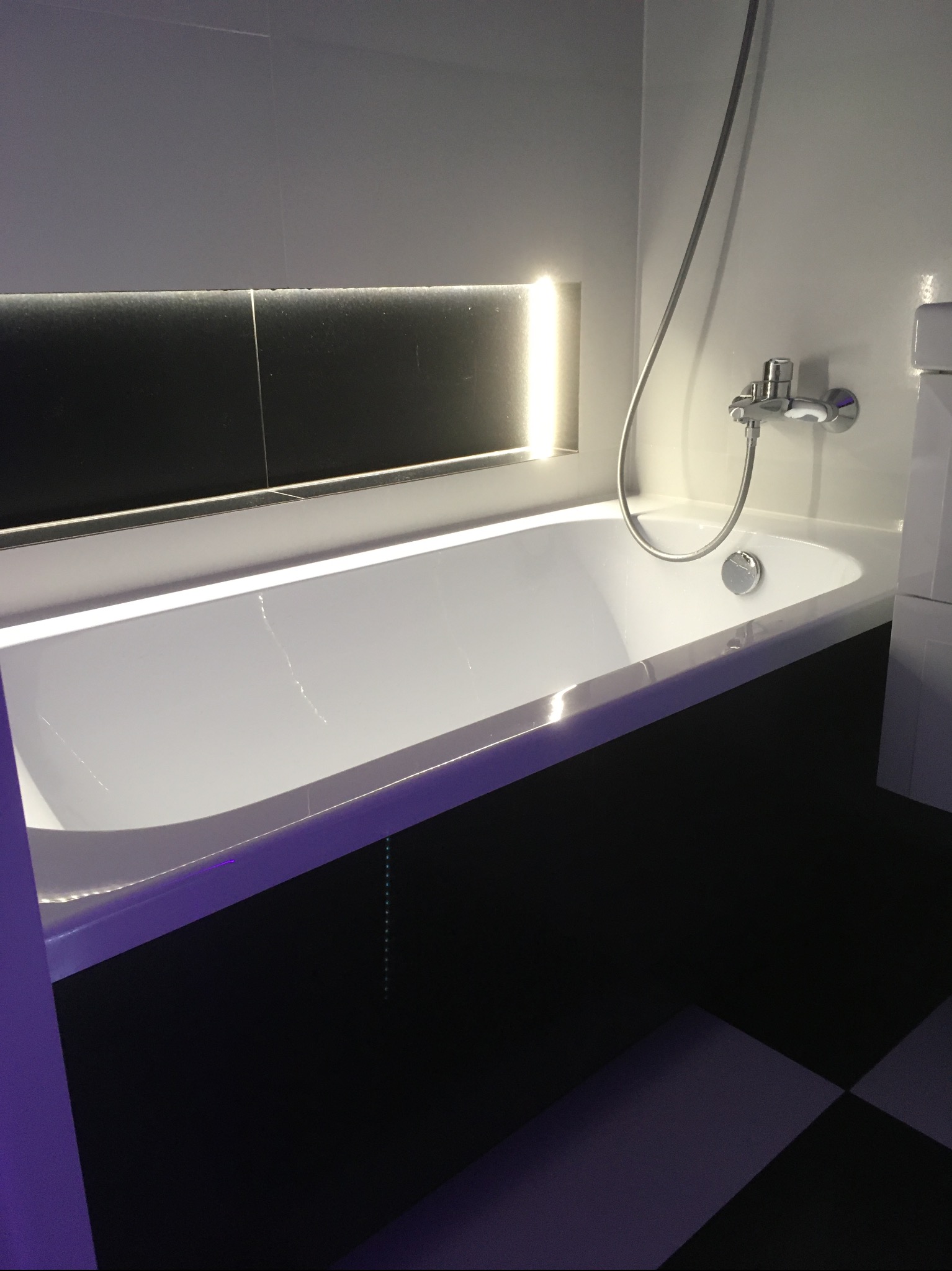 Nowoczesna, biała wanna z podświetleniem LED we wnęce, chromowana bateria, czarno-biała podłoga. Minimalistyczny design łazienki, eleganckie wykończenie.