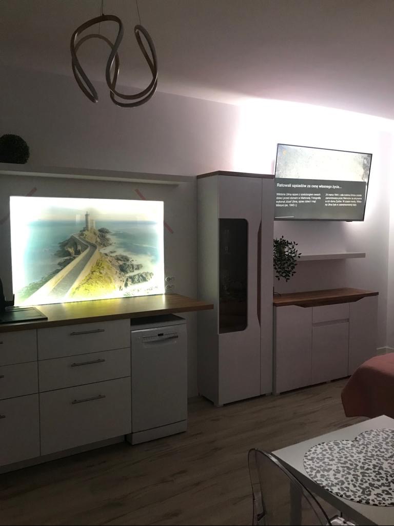 Nowoczesna kuchnia z białymi szafkami i blatem z drewna, oświetlona dekoracyjnym światłem. Na ścianie podświetlany obraz z motywem latarni morskiej i telewizor.