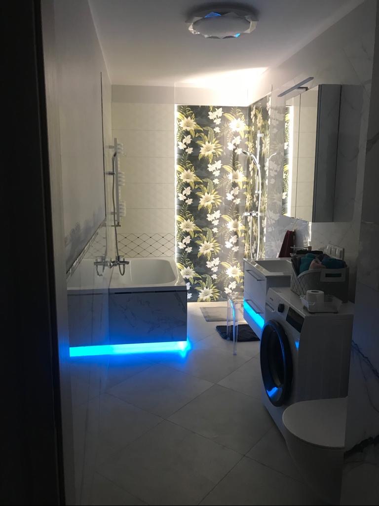 Nowoczesna łazienka z wanną podświetloną na niebiesko, kwiatową tapetą w kabinie prysznicowej i pralką. Minimalistyczny design, jasne płytki podłogowe.