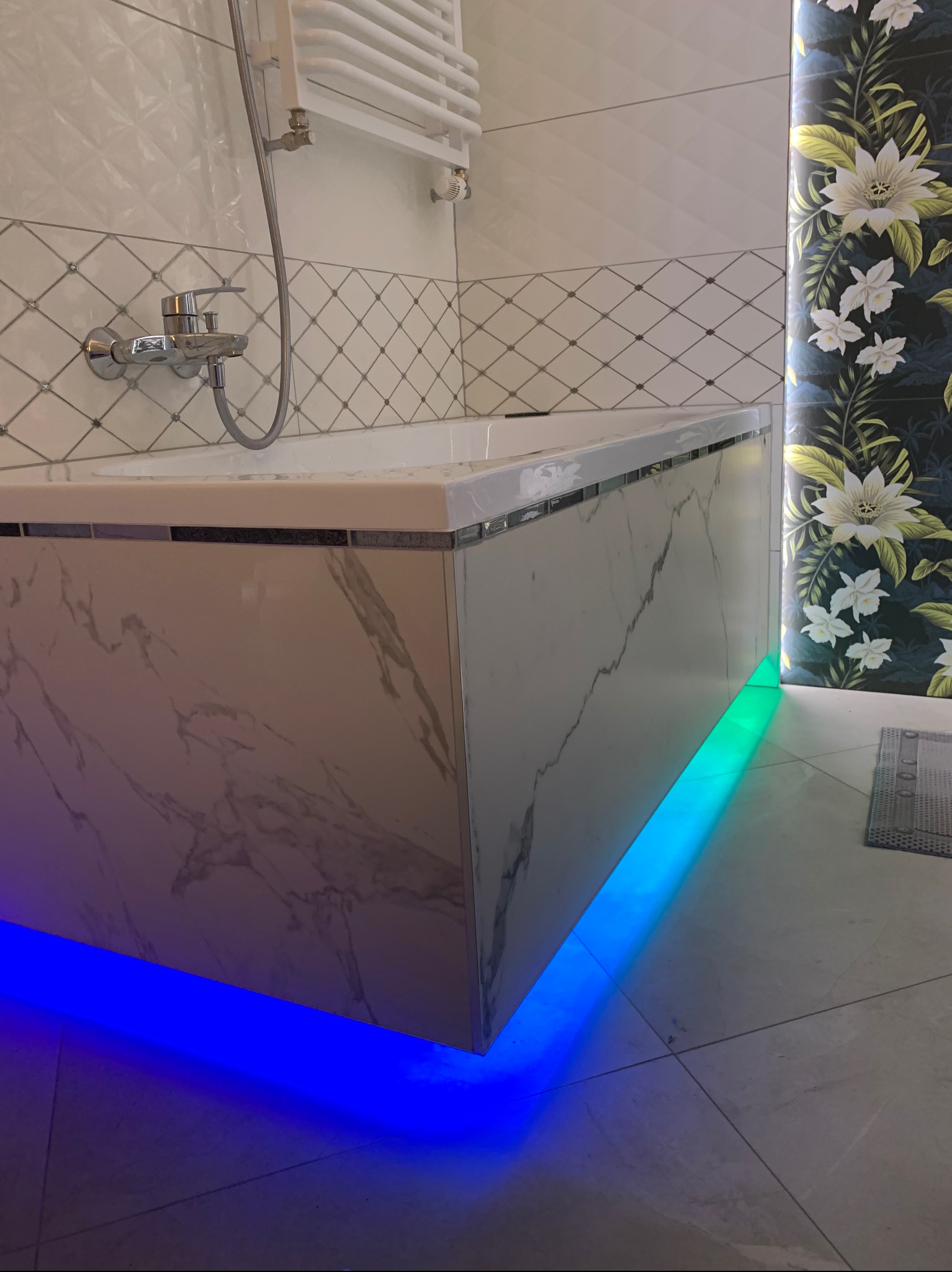 Wykończona wanna z marmurową obudową, podświetlona niebiesko-zielonymi LEDami, z baterią i grzejnikiem na tle płytek oraz dekoracyjnej tapety w kwiaty.