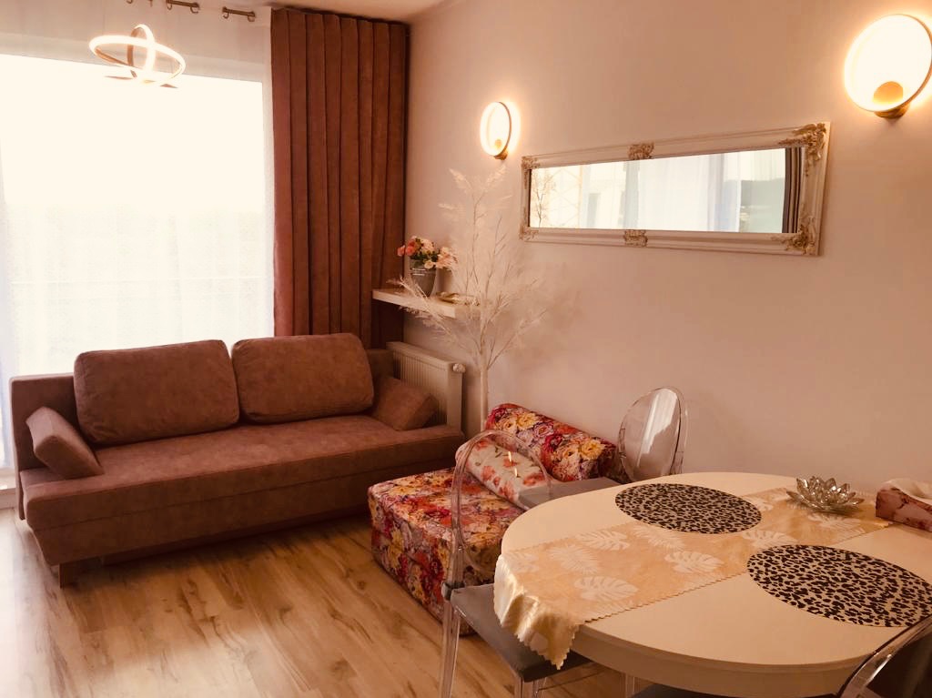 Jasne wnętrze salonu z brązową sofą, kwiatowym siedziskiem i okrągłym stołem. Dekoracyjne oświetlenie i lustro dodają elegancji. Widoczna drewniana podłoga.
