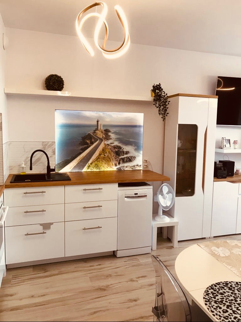 Biała, nowoczesna kuchnia z drewnianym blatem i motywem latarni morskiej na ścianie. Jasne wnętrze z designerską lampą i elementami dekoracyjnymi.
