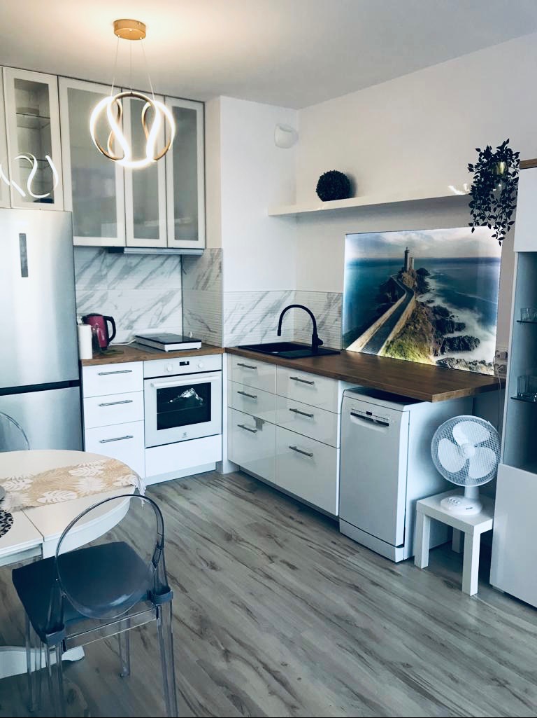 Jasna kuchnia z marmurowymi płytkami, okrągłym żyrandolem i stołem z transparentnymi krzesłami. Na blacie obraz z motywem latarni morskiej.