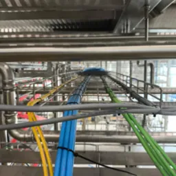 Kable w korytach kablowych, ułożone równolegle w różnych kolorach: żółtym, niebieskim, zielonym i szarym. Widok z dołu na instalację w hali przemysłowej.