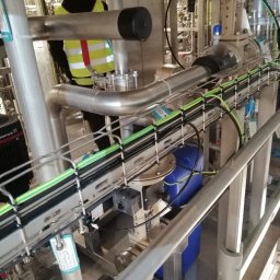 I-Markon - Przemysłowa instalacja z rurami, kablami w korytkach i pracownikiem w kasku. Widoczne metalowe elementy, zbiorniki i oznaczenia. Ujęcie z bliska, oświetlenie przemysłowe.