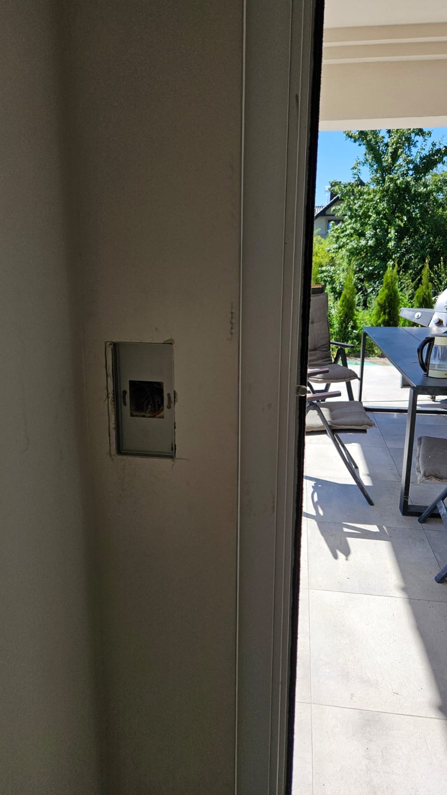 Puszka instalacyjna w ścianie, widoczna przez otwarte drzwi na taras z meblami ogrodowymi i zielenią. Przygotowanie do montażu gniazdka.