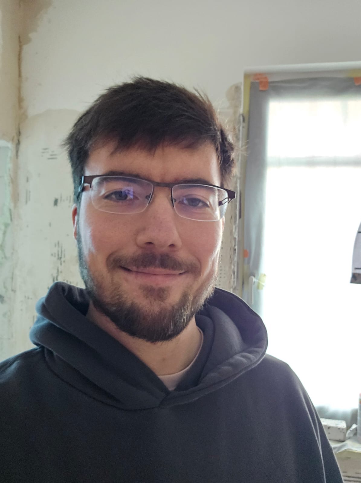 Uśmiechnięty mężczyzna w okularach i bluzie z kapturem na tle remontowanego pomieszczenia z oknem zabezpieczonym folią.