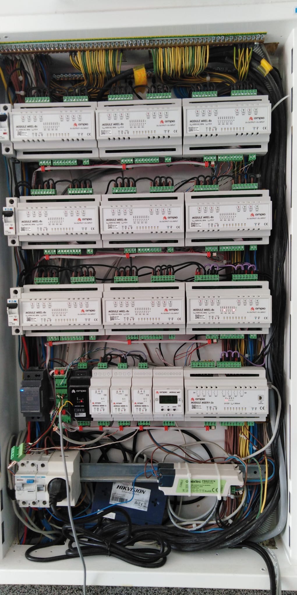 Rozdzielnia elektryczna z modułami Ampio MREL-8s, okablowaniem w kolorach żółtym, zielonym i czarnym, oraz urządzeniem Hikvision w dolnej części obudowy.