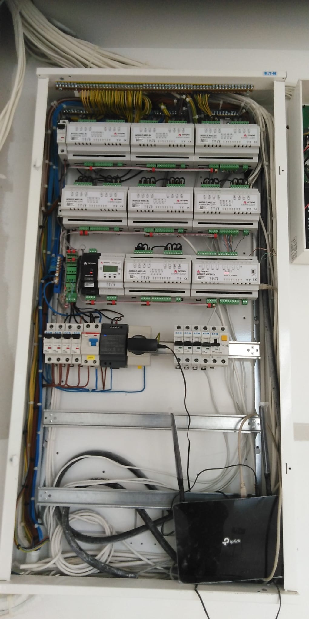 Rozdzielnica elektryczna z modułami, zabezpieczeniami i przewodami. Widoczny router TP-Link. Instalacja w skrzynce zasilającej, okablowanie wewnątrz szafki.
