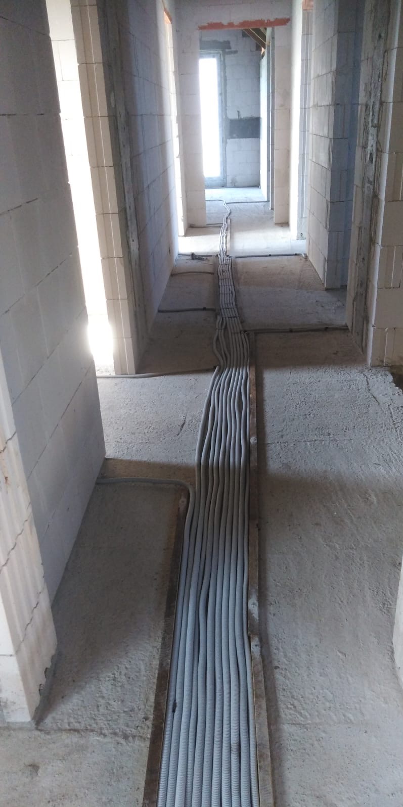 Szereg szarych rur instalacyjnych ułożonych równolegle w korytarzu budynku w trakcie budowy. Widoczne ściany z białych bloczków i surowy beton.