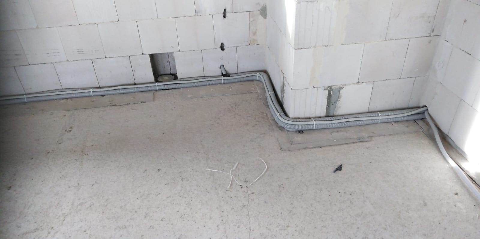 Szare rury instalacji elektrycznej prowadzone wzdłuż ściany z białych bloczków, zabezpieczone białymi opaskami zaciskowymi. Widoczne otwory w ścianie na kable.