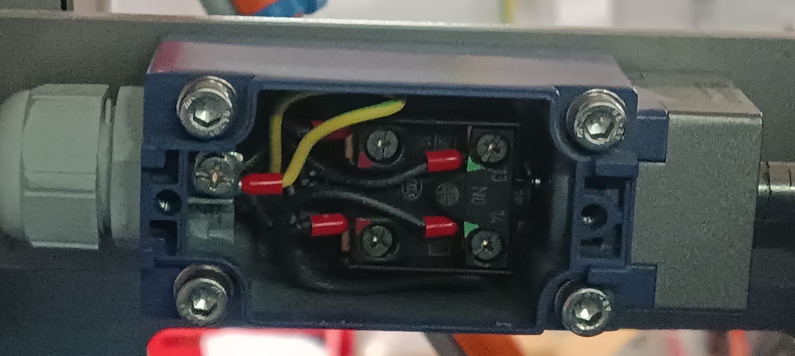 Szczegółowe ujęcie wnętrza puszki elektrycznej z przewodami w kolorach: żółto-zielonym, czarnym i czerwonym. Widoczne śruby mocujące i elementy złączne.