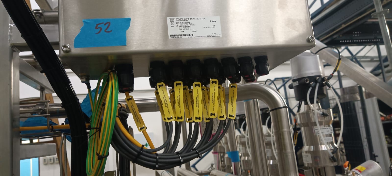 Metalowa skrzynka z numerem '52' i podłączonymi przewodami z żółtymi oznaczeniami, część instalacji przemysłowej, widoczne rury i elementy konstrukcyjne.