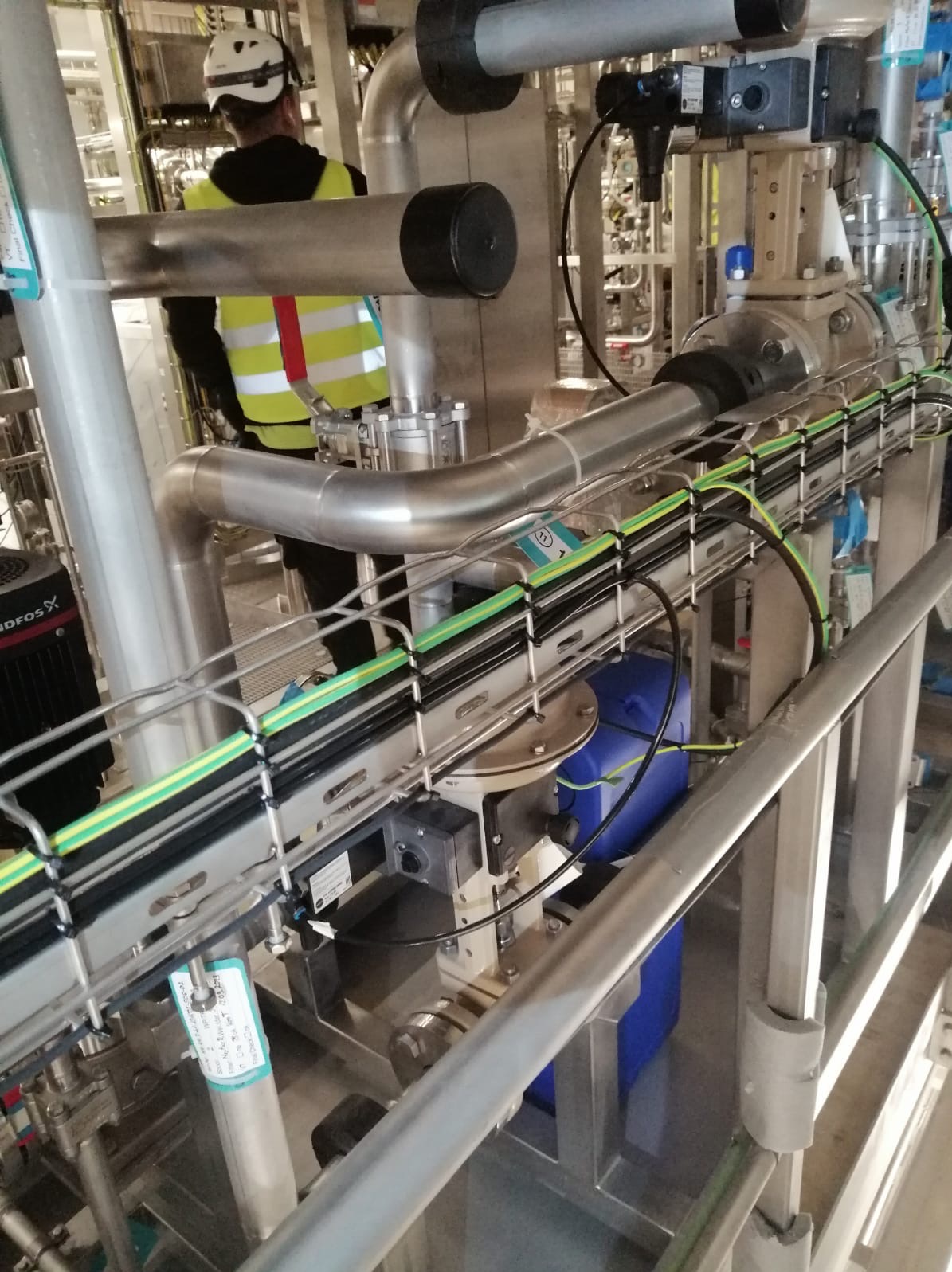 Przemysłowa instalacja z rurami, kablami w korytkach i pracownikiem w kasku. Widoczne metalowe elementy, zbiorniki i oznaczenia. Ujęcie z bliska, oświetlenie przemysłowe.