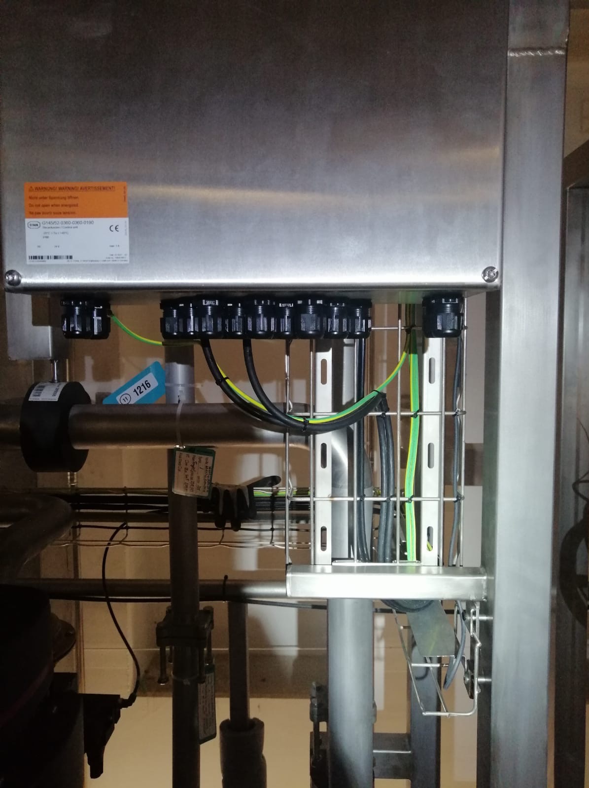 Metalowa skrzynka z podłączonymi przewodami elektrycznymi w osłonach, widoczne oznaczenia ostrzegawcze i tabliczka znamionowa. Przewody poprowadzone w metalowych korytkach.