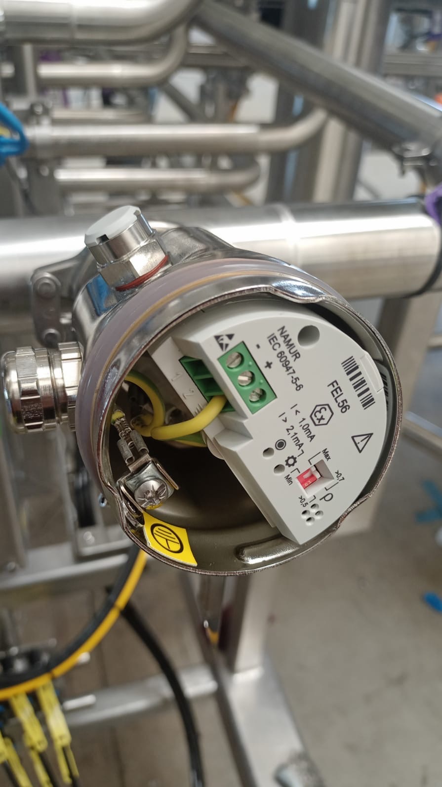 Otwarta obudowa urządzenia FEL56 z widocznymi złączami, okablowaniem i żółtą naklejką ostrzegawczą na tle metalowych rur. Widok z bliska na detale instalacji.
