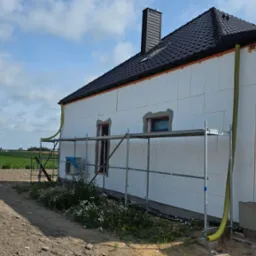 Dom w trakcie ocieplania: biały styropian na ścianach, rusztowanie, widok na pola i niebo. Etap prac z ż&oacute;łtymi rurami wentylacyjnymi. Dach pokryty czarną dach&oacute;wką.