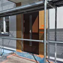 Elewacja domu w trakcie prac wykończeniowych: ocieplenie z drewnianymi panelami, widoczne rusztowania, okno i drzwi wejściowe. Częściowe zacienienie na ścianie.