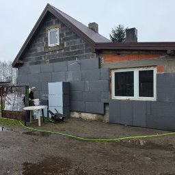 Tomasz Michałowski - Dom w trakcie ocieplania: szare płyty styropianowe na ścianach, widoczne okna, pracownik przy pracy. Częściowo ocieplona elewacja bloku z pustaków.