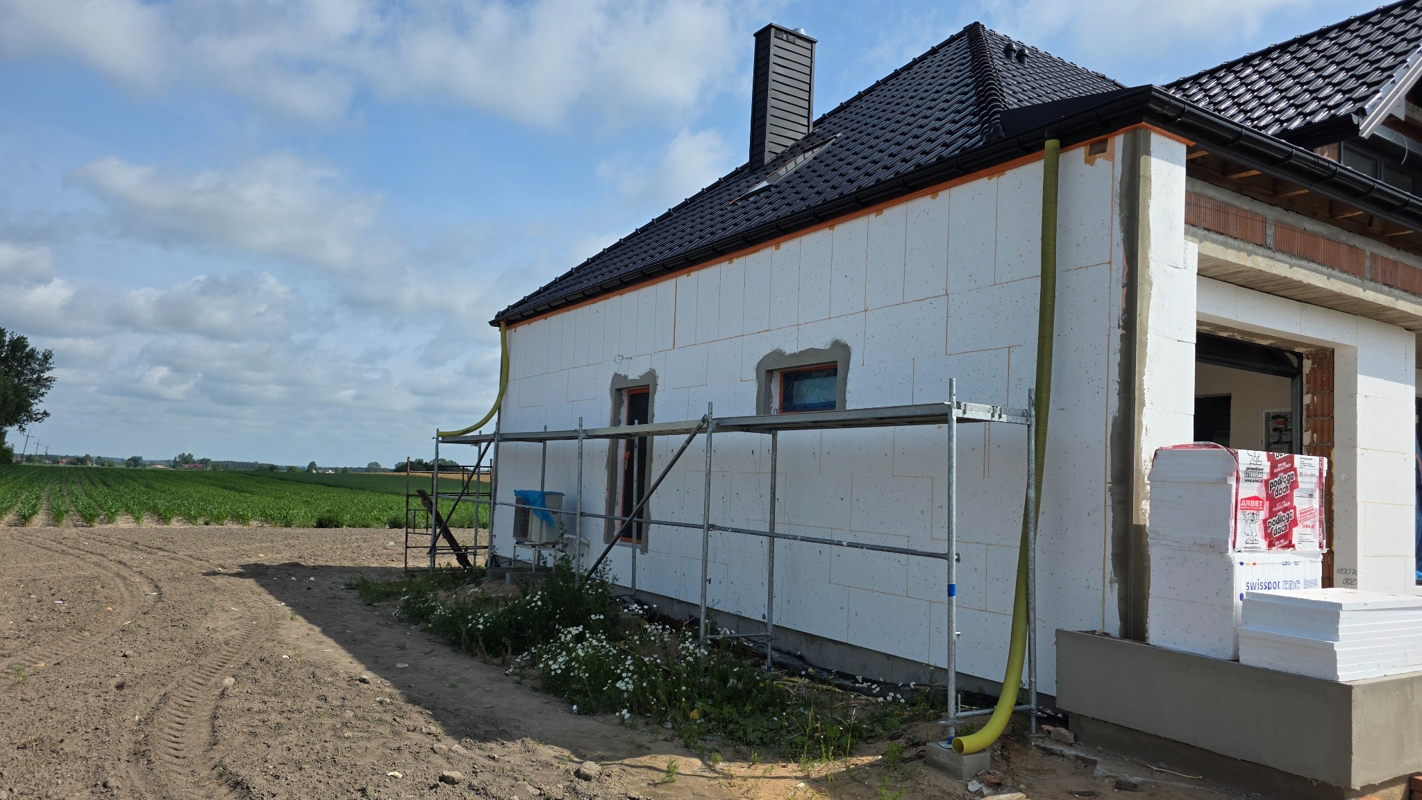 Dom w trakcie ocieplania: biały styropian na ścianach, rusztowanie, widok na pola i niebo. Etap prac z żółtymi rurami wentylacyjnymi. Dach pokryty czarną dachówką.