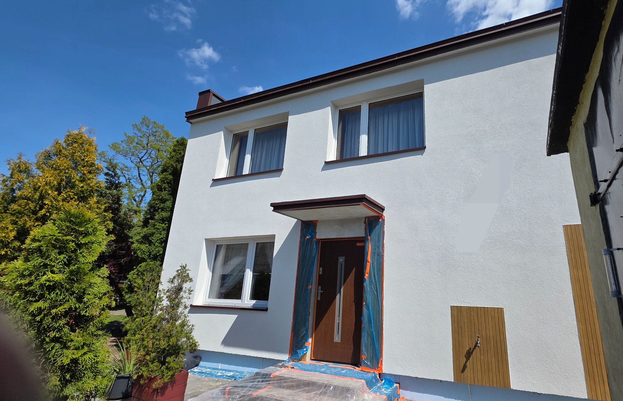 Biała elewacja domu jednorodzinnego z brązowym dachem i oknami. Drzwi wejściowe zabezpieczone folią malarską. Widoczne drzewa i błękitne niebo.