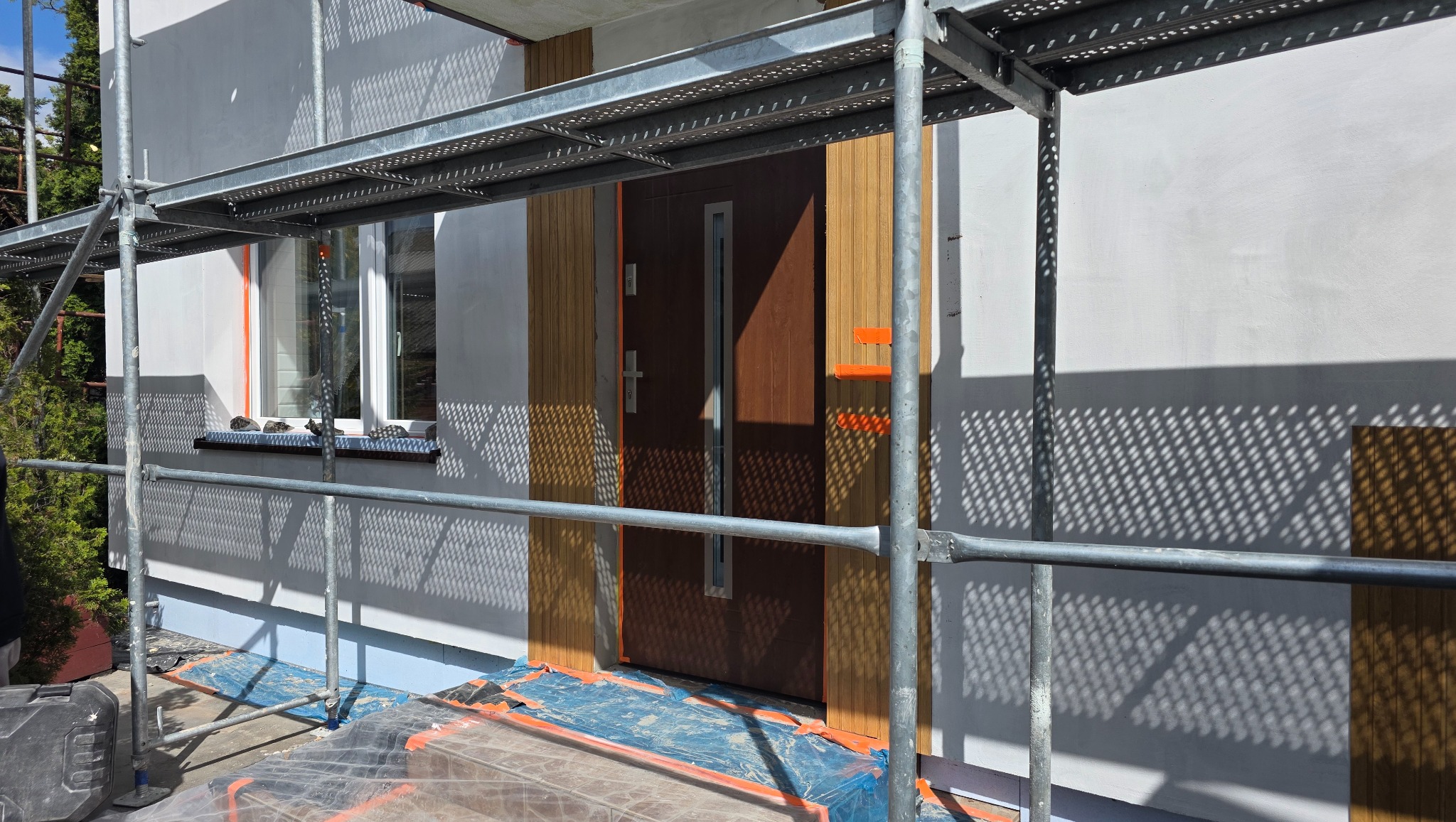 Elewacja domu w trakcie prac wykończeniowych: ocieplenie z drewnianymi panelami, widoczne rusztowania, okno i drzwi wejściowe. Częściowe zacienienie na ścianie.