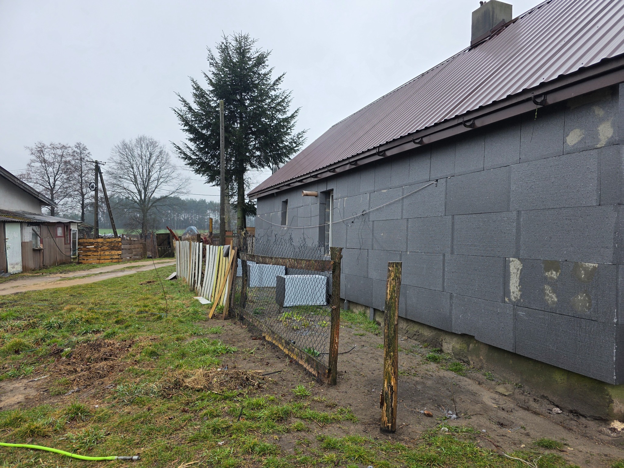 Szara elewacja domu w trakcie ocieplania, widoczne bloki styropianowe. W tle zielone drzewo iglaste i metalowy dach. Obok posesji płot z siatki.