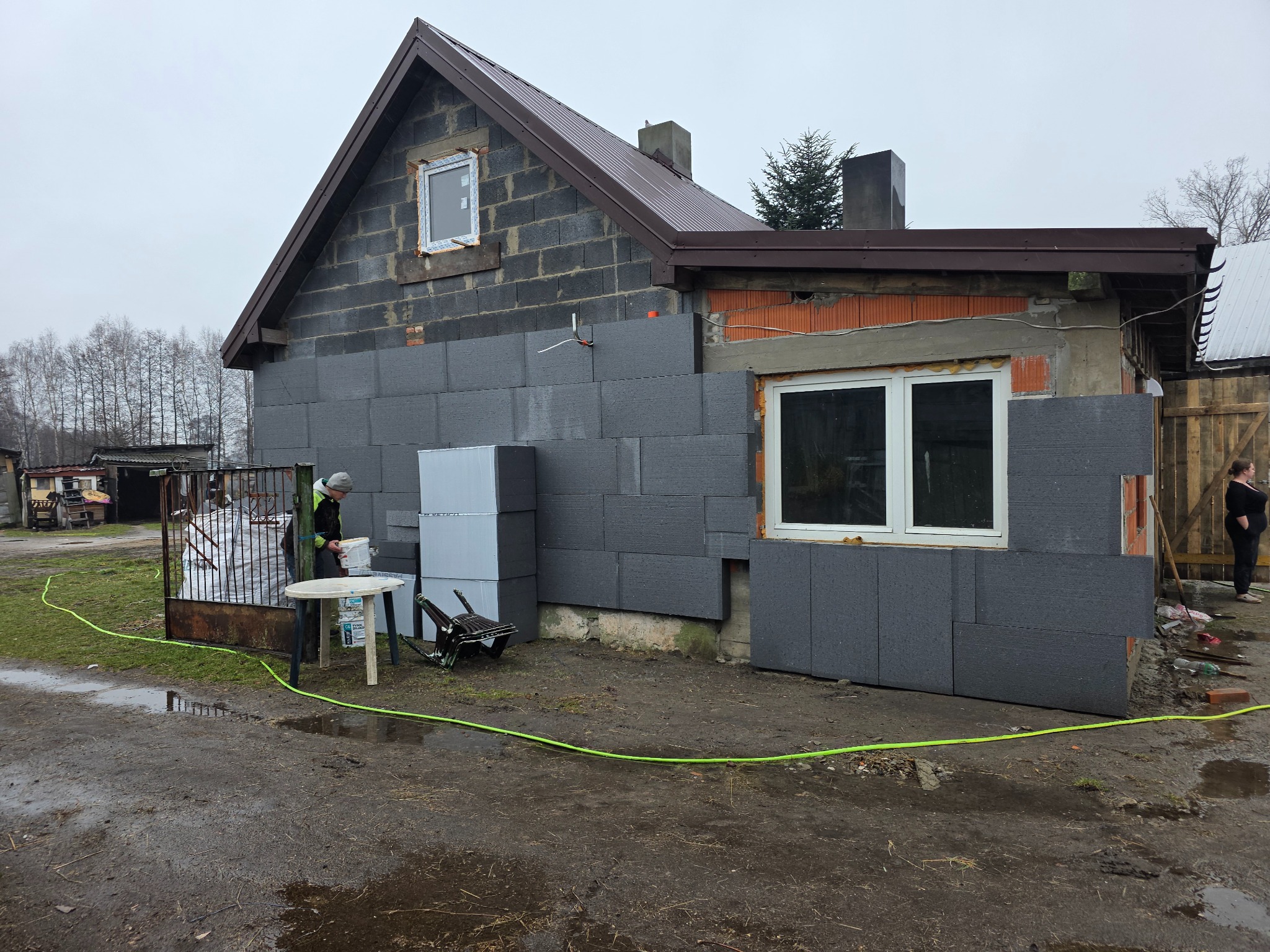 Dom w trakcie ocieplania: szare płyty styropianowe na ścianach, widoczne okna, pracownik przy pracy. Częściowo ocieplona elewacja bloku z pustaków.