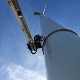 KASPOL Ireneusz Kaszowski - Praca na wysokości: Podnośnik koszowy przy turbinie wiatrowej na tle błękitnego nieba. Widoczny pracownik w koszu platformy.