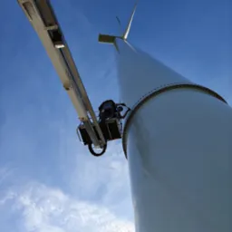 Praca na wysokości: Podnośnik koszowy przy turbinie wiatrowej na tle błękitnego nieba. Widoczny pracownik w koszu platformy.