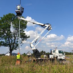 KASPOL Ireneusz Kaszowski - Pracownik w koszu podnośnika koszowego przy słupie oświetleniowym, drugi pracownik na ziemi w stroju roboczym. Biały podnośnik na tle błękitnego nieba.