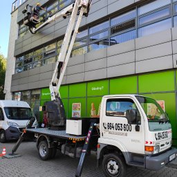 KASPOL Ireneusz Kaszowski - Pracownik w koszu podnośnika czyści okna biurowca. Biały podnośnik Kaspol na tle zielonej ściany. Widoczny numer telefonu. Praca na wysokości.