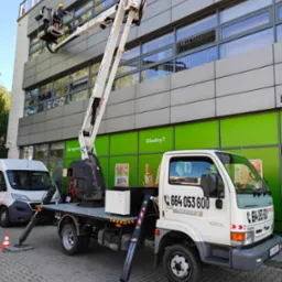 Pracownik w koszu podnośnika czyści okna biurowca. Biały podnośnik Kaspol na tle zielonej ściany. Widoczny numer telefonu. Praca na wysokości.