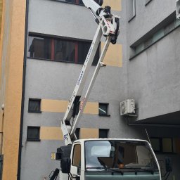 KASPOL Ireneusz Kaszowski - Podnośnik koszowy na tle budynku. Dwóch pracowników w pomarańczowych ubraniach w koszu podnośnika. Na pojeździe widoczny numer telefonu i napis 'WYNAJEM'.