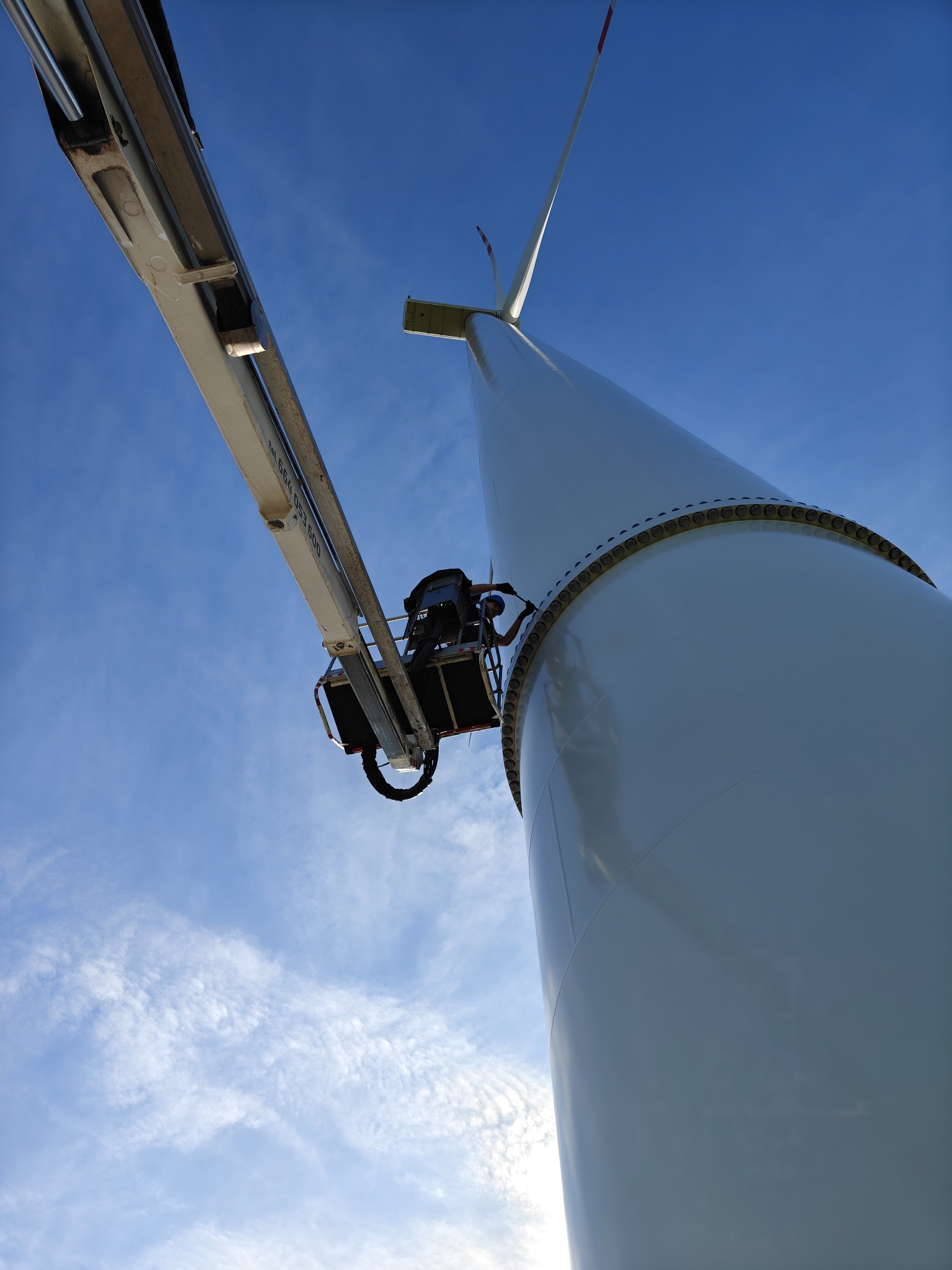 Praca na wysokości: Podnośnik koszowy przy turbinie wiatrowej na tle błękitnego nieba. Widoczny pracownik w koszu platformy.