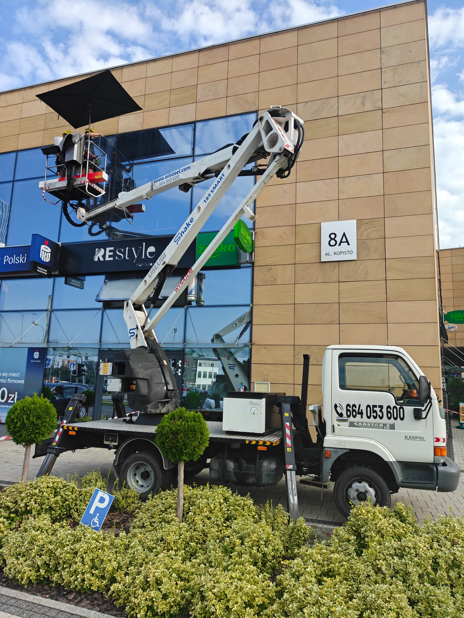 Podnośnik koszowy na tle budynku z przeszkloną fasadą podczas prac wysokościowych. Widoczny numer telefonu kontaktowego na kabinie pojazdu.