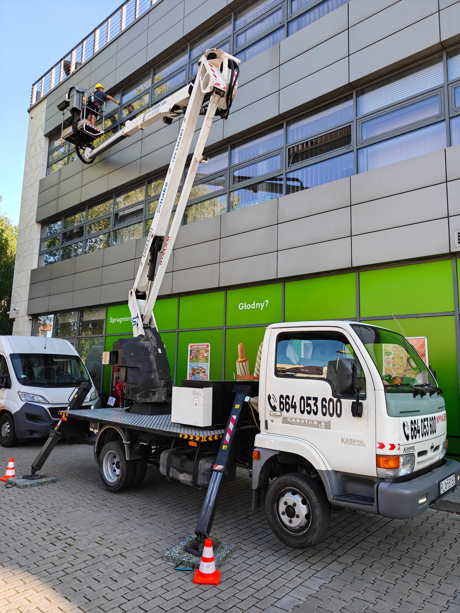 Pracownik w koszu podnośnika czyści okna biurowca. Biały podnośnik Kaspol na tle zielonej ściany. Widoczny numer telefonu. Praca na wysokości.