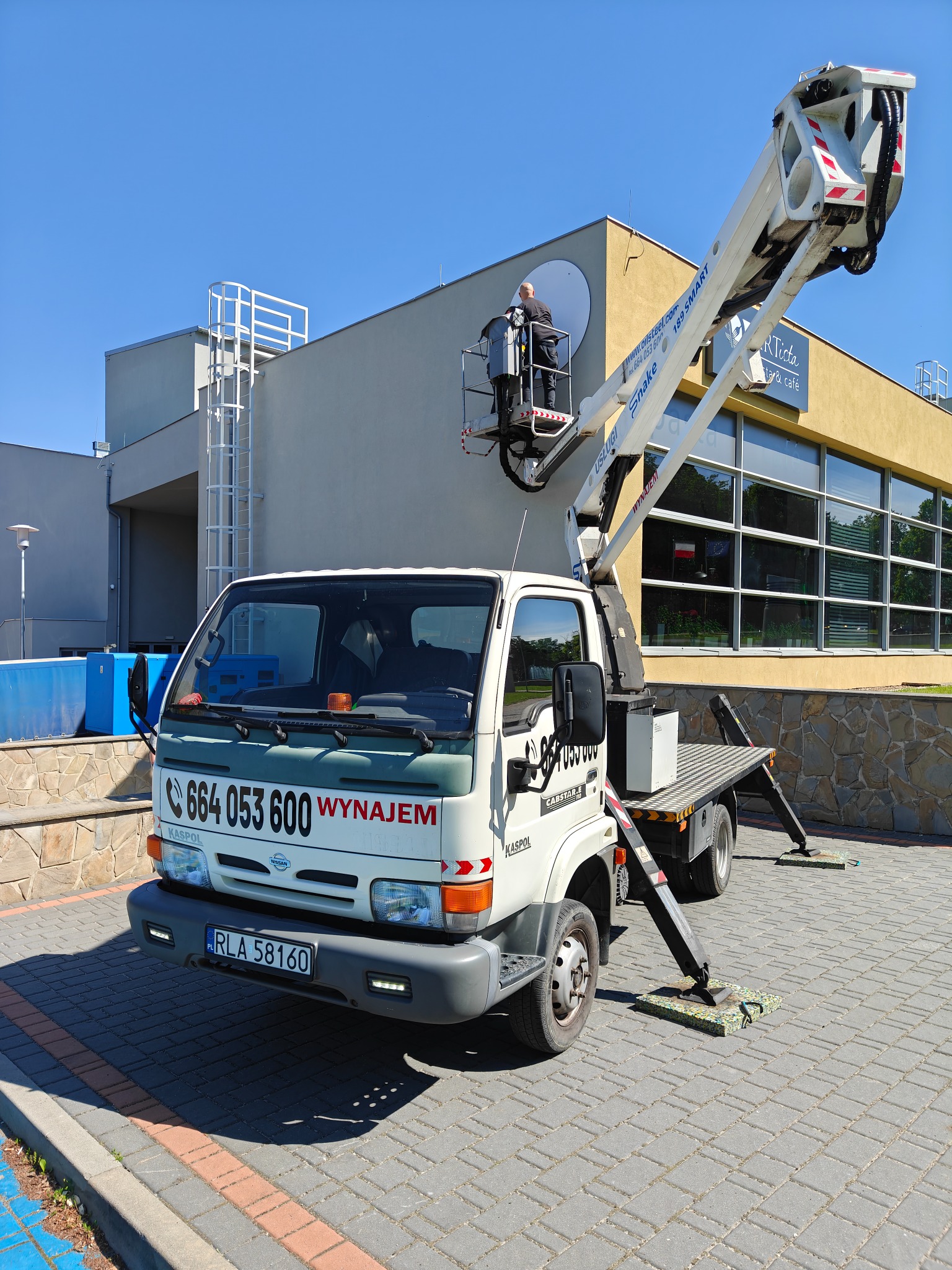 Podnośnik koszowy KASPOL na tle budynku z szyldem restauracji, pracownik w koszu na wysokości, mycie okien lub montaż reklamy, błękitne niebo.